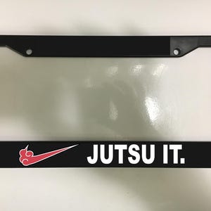 以下が含まれることがあります： 白い文字で「JUTSU IT.」と書かれた黒いナンバープレートフレーム。フレームには、スウッシュロゴに似た赤と白のデザインが施されています。フレームは角が丸い長方形です。