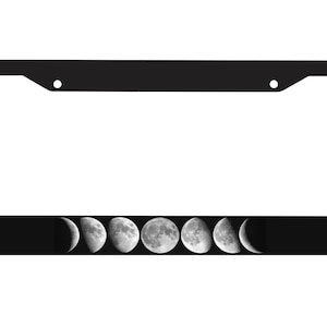 May include: Black license plate frame featuring a series of moon phases across the bottom. The phases progress from a crescent to a full moon and back, creating a celestial design. The frame has a rectangular shape with rounded corners.