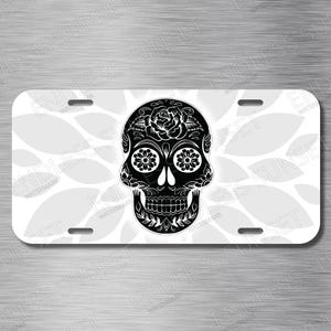 May include: A decorative license plate featuring a detailed black sugar skull design on a white background. The skull is adorned with floral patterns and intricate details. The background has a subtle pattern of gray leaves.