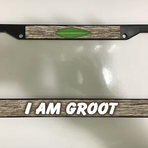 May include: Black license plate frame with a wood grain pattern and a green leaf accent. The phrase "I AM GROOT" is displayed in white, bold letters across the bottom. The frame is designed to hold a standard license plate.