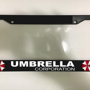 May include: Black license plate frame with the text "UMBRELLA CORPORATION" in white. The frame has a black border and features a red and white logo on each side of the text.