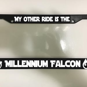 May include: Black license plate frame with white text that reads "MY OTHER RIDE IS THE MILLENNIUM FALCON." The frame has a Star Wars logo on each side. The frame is rectangular and fits a standard U.S. license plate.