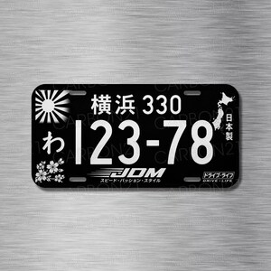 May include: A black rectangular license plate featuring white text and graphics. It displays the Japanese characters for Yokohama, the number 330, and 123-78. It includes a rising sun emblem, a map of Japan, cherry blossoms, and the text JDM Drive Life.