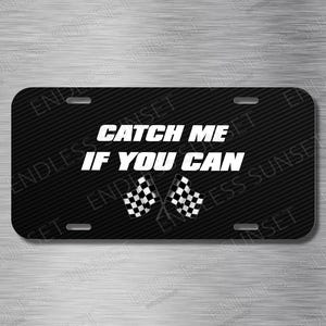 May include: Black license plate with the text "CATCH ME IF YOU CAN" in white. Two checkered racing flags are below the text. The plate has a carbon fiber texture and four mounting holes.
