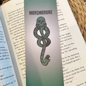 May include: A green bookmark with a dark green snake-like symbol and a skull. The symbol is called a "Morsmordre" and is a dark magic symbol. The bookmark is on a book with text that reads "Put down your sword".