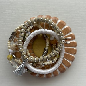 May include: A collection of beaded bracelets in neutral tones, including white, beige, and silver, are arranged on a decorative, circular, tan-colored base. The bracelets feature various bead sizes and textures, with a silver tassel adding a touch of elegance.