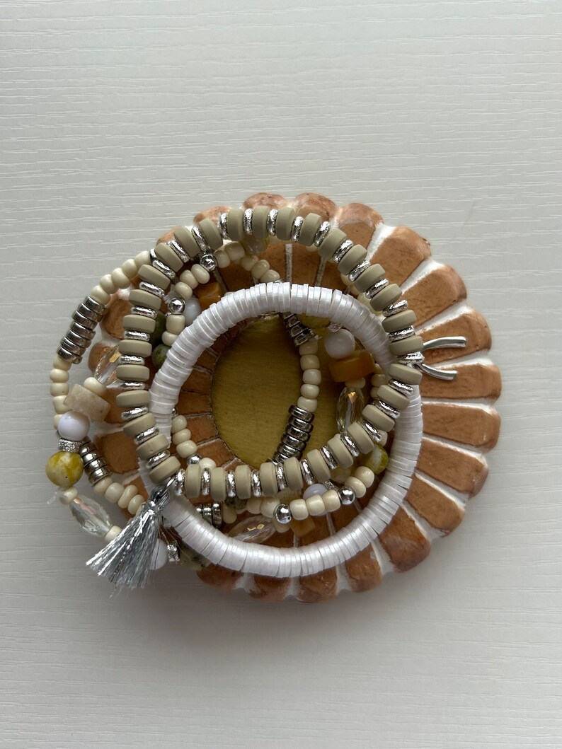 May include: A collection of beaded bracelets in neutral tones, including white, beige, and silver, are arranged on a decorative, circular, tan-colored base. The bracelets feature various bead sizes and textures, with a silver tassel adding a touch of elegance.