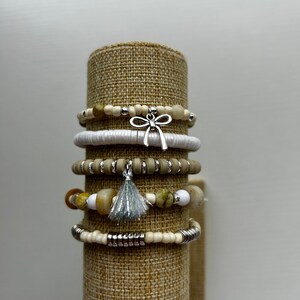 May include: Stack of beaded bracelets in neutral tones, displayed on a cylindrical stand. The bracelets feature a variety of beads, including white, tan, and stone-like textures, accented with silver-toned metal and charms, including a bow and a tassel.