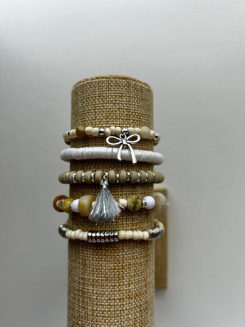May include: Stack of beaded bracelets in neutral tones, displayed on a cylindrical stand. The bracelets feature a variety of beads, including white, tan, and stone-like textures, accented with silver-toned metal and charms, including a bow and a tassel.