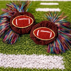 May include: Two brown hair accessories, each featuring a football design with a white and gold outline. The accessories have a textured appearance and are adorned with colorful, iridescent fringe, set against a green background.