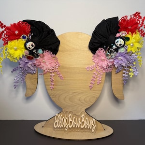 May include: Two black fabric hair bows adorned with red, yellow, lavender, and pink artificial flowers, pearl accents, and small skeleton figurines. The bows are displayed on a wooden stand featuring the text Ella's Bow-Bows in white script.