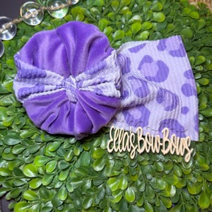 May include: A purple baby headband with a velvet bow and a matching leopard print band. The bow is a deep purple, and the band features a lighter purple leopard print. The headband is displayed on a bed of green leaves.