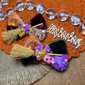 May include: Two hair bows featuring velvet and sequin fabric in orange, purple, and black. Each bow is decorated with a miniature straw broomstick and a small purple or gold sequin band. A clear acrylic sign with the text Ella's Bow Bows is included.