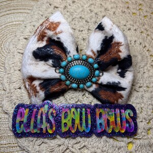 May include: A cow print hair bow with a turquoise and silver embellishment. The bow is paired with a purple glittery barrette that reads "ELLA'S BOW BOUTIQUE" in colorful letters. The bow is set on a beige crocheted surface.