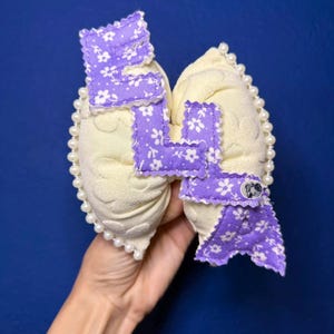 May include: A handmade hair bow featuring a cream-colored base with a floral pattern and a purple floral letter "L" applique. The bow is trimmed with small white pearls and has a small button detail.