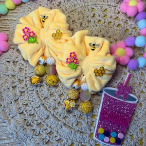 May include: Yellow scrunchies with bee and flower embellishments, alongside beaded accents. A decorative purple glitter drink charm and a pastel pom-pom garland are also present. The items are arranged on a beige crocheted surface.