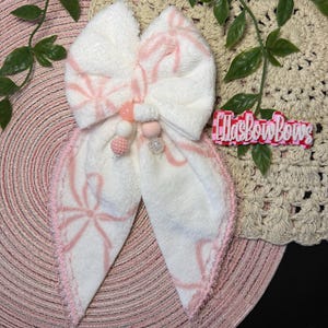 May include: A large white bow with pink accents and a beaded center. The bow features a pink bow pattern and a pink beaded trim. The bow is on a pink circular mat with a beige crocheted background and the text "EllasBowBows".