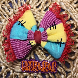 May include: A fabric hair bow featuring a patchwork design of yellow, bright blue, and magenta ribbed fabric sections. The bow is accented with black stitched details and a red lace trim along the edges. Below the bow is a purple glitter sign reading EllasBowBows.