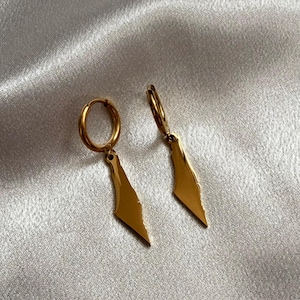 May include: A pair of gold-colored hoop earrings. Each earring features a small hoop with a geometric, elongated teardrop-shaped pendant. The earrings are displayed on a shiny, light-colored fabric.