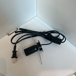 May include: A black light fitting with a cable and plug. The fitting has a black socket and a metal bracket. The cable is black and has a switch. The plug is white and black. The fitting is on a white surface.