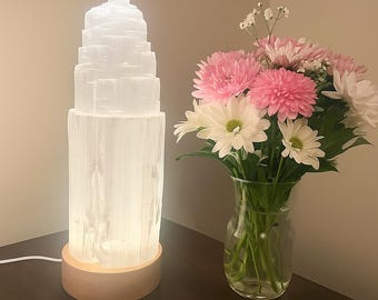 Könnte beinhalten: Eine weiße Selenit-Turmlampe mit Holzsockel leuchtet und strahlt ein sanftes Licht aus. Ein Strauß rosa und weißer Blumen in einer Glasvase steht daneben. Das Design der Lampe und die Blumen schaffen eine ruhige Atmosphäre.