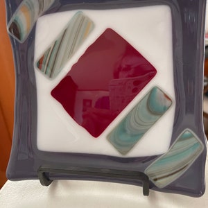 May include: A square glass dish with a purple border and a white center. The center features a diamond shape made of red, white, and blue glass.