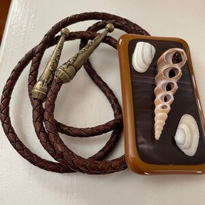 Op de afbeelding: Een bruine leren bolo tie met een rechthoekige glazen hanger met een schelpontwerp. De hanger heeft een bruine achtergrond met een witte schelp en een bruine schelp met een spiraalvormig patroon.