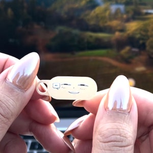 May include: A small, beige, rectangular charm with a cartoon face and a speech bubble. The charm has a small hole at the top for attaching to a keychain or bracelet.