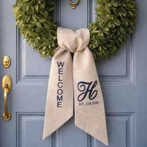 May include: A green wreath hangs on a light blue door, adorned with a beige ribbon. The ribbon features the word "WELCOME" in navy blue lettering on one side and a navy blue monogram with the date "EST. 2.24.2026" on the other.