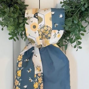 May include: A decorative fabric item with a patchwork design featuring sunflowers, bees, and honeycomb patterns. The item has a blue and yellow color scheme and is tied in a knot. The item is hanging in front of a green wreath.