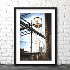 May include: A framed photograph featuring a tall, cylindrical water tower with the text Buffalo Trace and a buffalo graphic. The tower stands above industrial brick buildings and metal pipes under a cloudy sky, captured in a vertical composition.