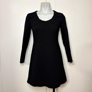 May include: A solid black long-sleeve dress featuring a scoop neckline and structured, pleated detailing along the upper chest and shoulder area. The garment has a fitted bodice that flares slightly toward the hem, creating a simple silhouette.