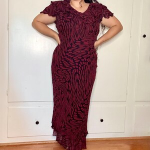 May include: A burgundy formal dress with a black pattern and a V-neck. The dress has short, ruffled sleeves and a flowing skirt. The dress is floor-length and has a subtle shimmer.