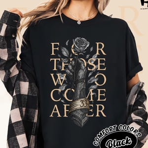 Puede incluir: Camiseta negra con estampado gráfico con el texto "FOR THOSE WHO COME AFTER" y una ilustración de rosa. La camiseta también presenta el número 33 y las palabras "COMFORT COLORS Black". La modelo lleva una camisa a cuadros negra y beige.