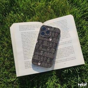 May include: A black phone case with a bookshelf design featuring books, bottles, and floral elements in white and pink. The case is resting on an open book on a bed of green grass.