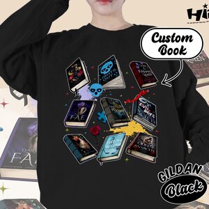 May include: Black sweatshirt featuring a collection of illustrated books with titles like "Lights Out" and "Evil Boys". The design includes a skull, puzzle pieces, and a rose. The text "Custom Book" and "GILDAN Black" are also visible.