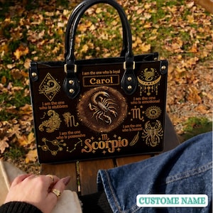 Puede incluir: Un bolso negro con gráficos dorados del signo zodiacal Escorpio, que incluye un escorpión central, constelaciones y el texto Carol, Scorpio, además de frases como I am the one who is loyal, stubborn, emotional, possessive, and honest.