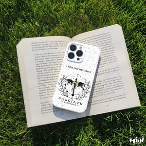 May include: White speckled phone case with the text "I WILL NOT DIE TODAY" and a black and gold graphic. The case is lying on an open book on a bed of green grass.