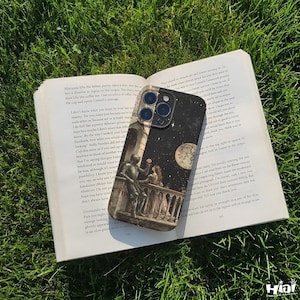 May include: A phone case featuring a balcony scene under a starry night sky and a full moon. The case is placed on an open book on green grass. The book has text on the pages.