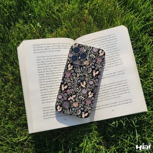 Może przedstawiać: Czarny pokrowiec na telefon z kwiatowym wzorem z ptakami i kwiatami w odcieniach różu, beżu i złota. Etui leży na otwartej książce na zielonej trawie. Etui ma błyszczące wykończenie.