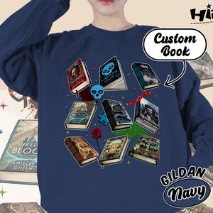 May include: Navy blue sweatshirt featuring a collection of illustrated books with titles like "Lights Out" and "Promise Peridot." The design includes a skull, a rose, and the text "Custom Book." The sweatshirt is labeled "GILDAN Navy."