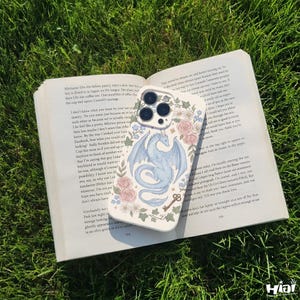 May include: A white phone case with a floral design and a light blue dragon illustration. The case is resting on an open book on a bed of green grass. A small key graphic is also visible.