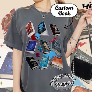 May include: A gray Comfort Colors t-shirt featuring a collection of illustrated books with titles like "Lights Out" and "Academy of Liars." The shirt also has the text "Custom Book" and "Pepper."
