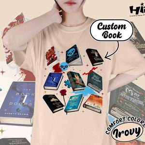 May include: Beige t-shirt featuring a collection of illustrated books with various covers, including titles like "Lights Out" and "The Book of 7 Doors." The design includes a skull, a rose, and the text "Custom Book."