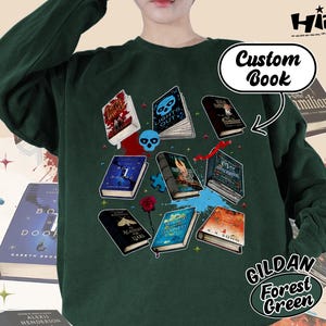 May include: Forest green sweatshirt featuring a collection of illustrated books with titles like "Lights Out" and "Academy for Liars." The design includes a skull, puzzle pieces, and a rose. The text "Custom Book" is also visible.