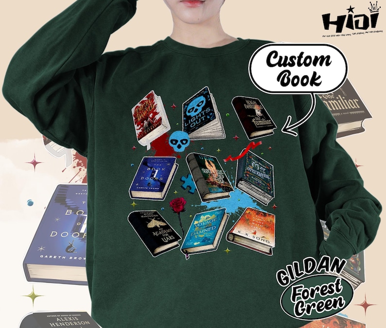 May include: Forest green sweatshirt featuring a collection of illustrated books with titles like "Lights Out" and "Academy for Liars." The design includes a skull, puzzle pieces, and a rose. The text "Custom Book" is also visible.