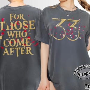 May include: Dark grey t-shirts with gold text and red accents. One shirt reads "FOR THOSE WHO COME AFTER" and the other displays the number "33". The shirts are from Comfort Colors.