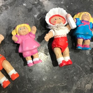 May include: A collection of six Cabbage Patch Kid dolls. The dolls are all wearing different outfits and have different hair colors. The dolls are all smiling and have big eyes.