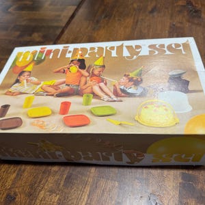 May include: A vintage "mini party set" box with a colorful illustration of children at a party. The box features various party supplies, including plates, cups, and a cake. The box is on a wooden surface.