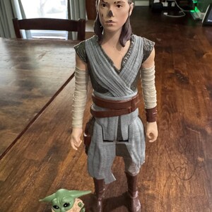 K&ouml;nnte beinhalten: Eine gro&szlig;e Actionfigur eines Charakters aus dem Star Wars-Franchise, die ein grau-blaues Outfit mit braunen Stiefeln und einem G&uuml;rtel tr&auml;gt. Eine kleine gr&uuml;ne Figur von Grogu befindet sich zu F&uuml;&szlig;en der Figur. Die Figuren stehen auf einer Holzoberfl&auml;che.